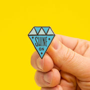 diamond shaped enamel pin that says "shine on" in hand