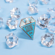 diamond shaped enamel pin that says "shine on" on blue background