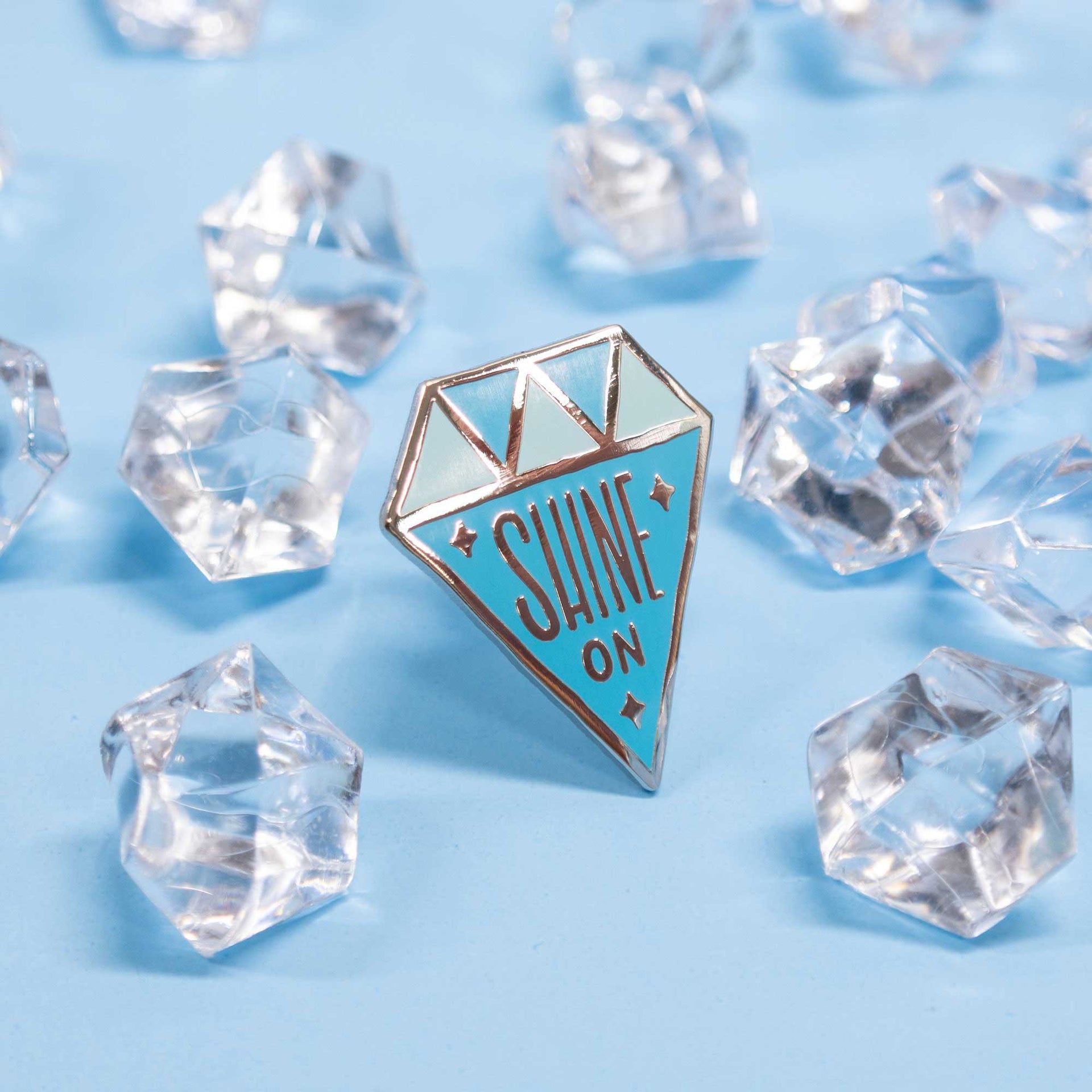 diamond shaped enamel pin that says "shine on" on blue background