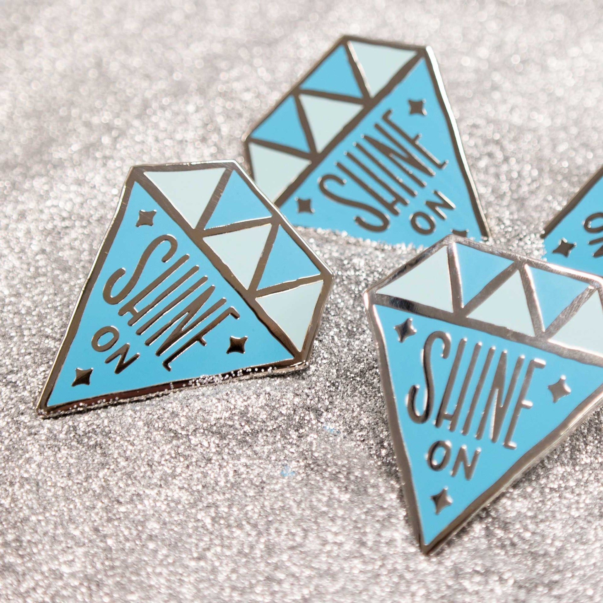 diamond shaped enamel pins that says "shine on" in silver glitter
